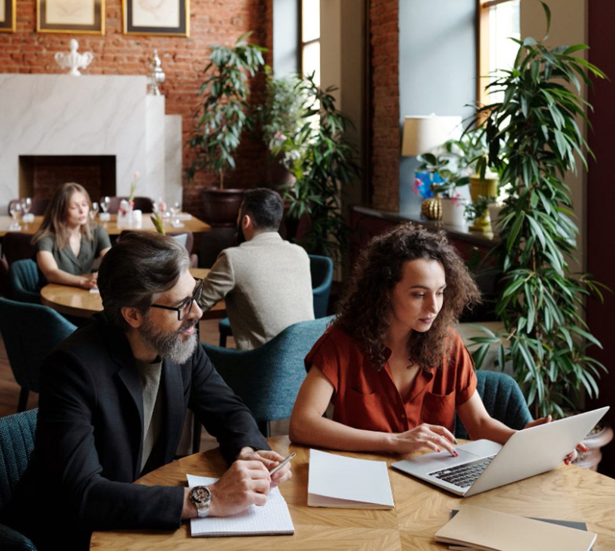 Two colleagues reviewing work together on a laptop in a warm, plant-filled café workspace